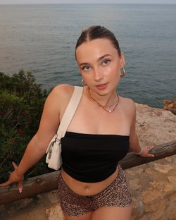 A young woman with brown hair and blue eyes wears a black crop top and leopard-print shorts, posing by the ocean on a