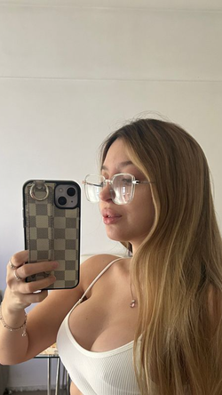 Curvy woman with long blonde hair and glasses takes a selfie in a white tank top indoors.