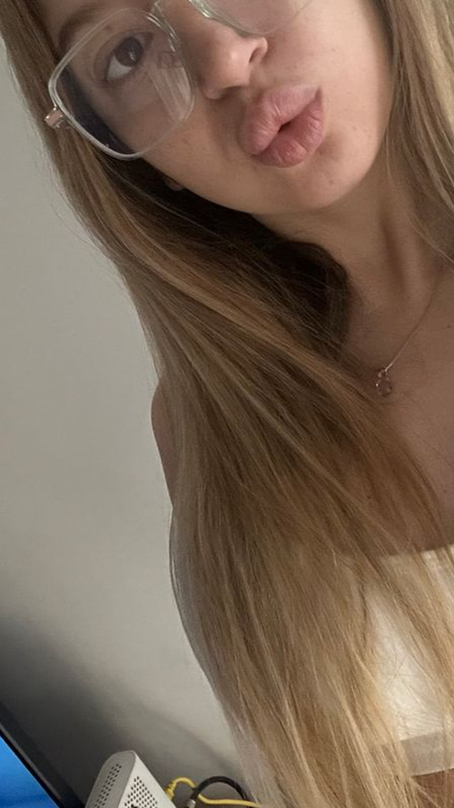 Slim blonde woman with glasses and long hair puckers her lips in a selfie.