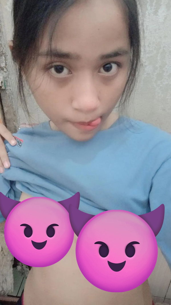 A young woman with dark hair in a blue shirt poses in a selfie, sticking her tongue out with emoji covering her chest.