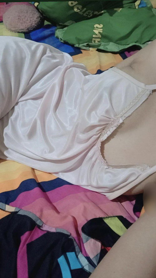 Slim woman in white satin slip on colorful messy bed, side profile with slipped strap.