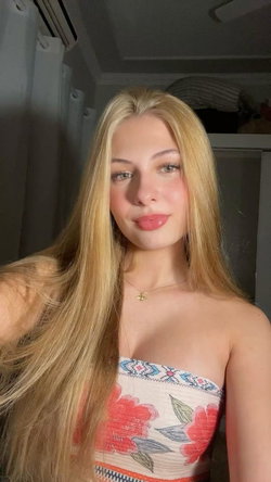 Blonde woman in a floral strapless top poses for a selfie in a dimly lit room.