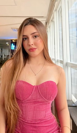 A woman with long blonde hair and a pink strapless dress stands indoors near a window.
