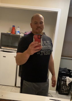 Bald man with beard taking mirror selfie in black t-shirt and gray pants, standing in a kitchen with appliances and