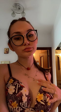Woman with glasses in a floral top takes a selfie indoors with hand on her chest.