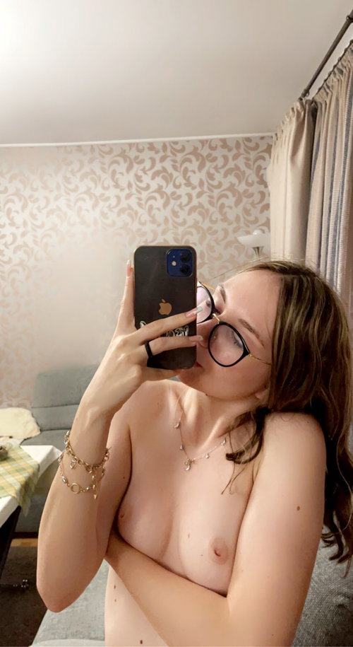 Brunette woman with glasses takes a topless selfie in a bedroom setting.