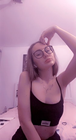 Slim woman with glasses wearing a black crop top posing in a bedroom under purple light.
