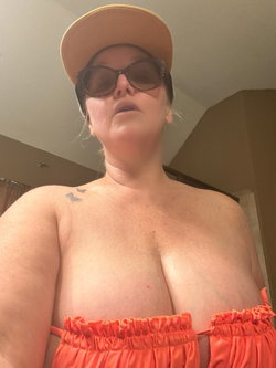 Curvy woman wearing a beige cap and sunglasses, topless with orange ruffled bottoms, indoors.