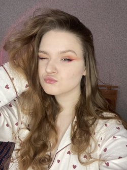 A young woman with long wavy brown hair and a nose ring, wearing heart-patterned pajamas, posing indoors with one eye