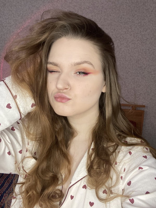 A young woman with long wavy brown hair and a nose ring, wearing heart-patterned pajamas, posing indoors with one eye