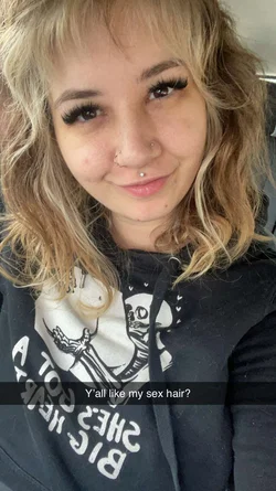 Blonde woman with nose and lip piercings smiles wearing graphic black hoodie in car with Snapchat overlay.
