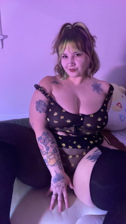 Curvy tattooed woman in sheer lemon-print top and black pants sits on bed in purple-lit room.
