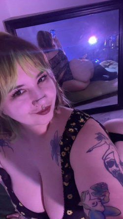 Curvy woman with colorful hair and tattoos takes a mirror selfie in a dimly lit room.