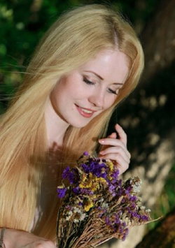 Blonde woman with long hair smiles softly while holding a bouquet of dried flowers in a wooded setting.