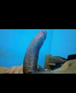 Close-up of an erect penis with hands visible, set against a blue wall and bed in a dimly lit room.