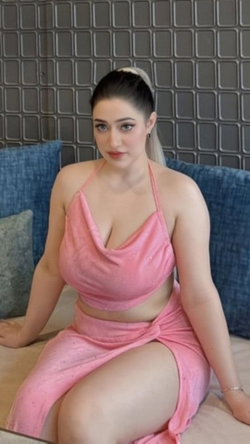 Curvy woman in a pink velour two-piece outfit sits on a bed with blue pillows, hair in a high ponytail, looking at the