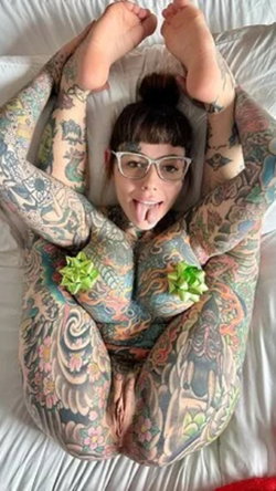 Tattooed woman with dark hair and glasses lying on a bed, legs raised, sticking tongue out playfully.