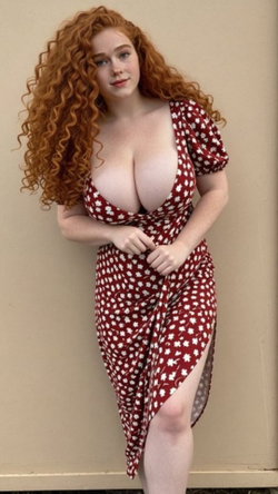 A curvy redhead with long curly hair stands in a red floral dress against a plain background.