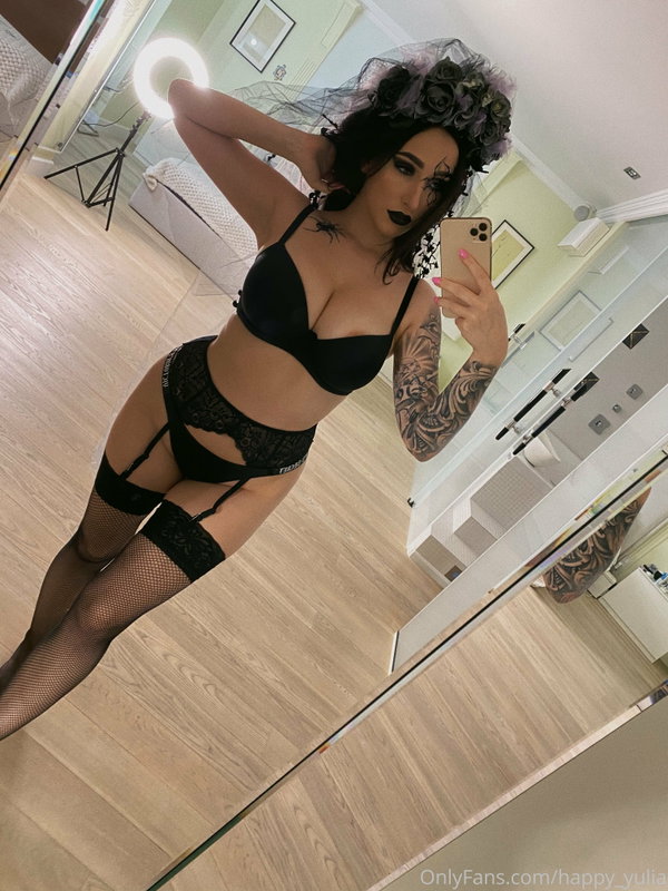 A woman with dark hair and dramatic makeup takes a mirror selfie in black lingerie, fishnet stockings, and a floral