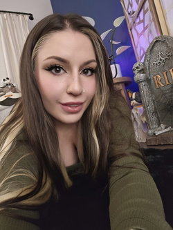 Slim woman with brunette hair and blonde highlights wears green sweater in Halloween room.