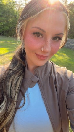 Smiling woman with blonde ponytail, freckles, nose piercing outdoors in blue top and tan jacket.