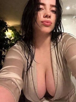 Curvy woman with long wet dark hair wearing a sheer low-cut top takes a selfie indoors.