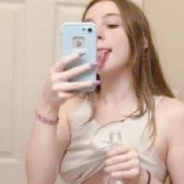 A woman with long light brown hair takes a mirror selfie in a light-colored tank top, sticking her tongue out while