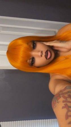 Woman with vibrant orange hair and bold makeup poses indoors, hand near face, visible arm tattoo and nose piercing.