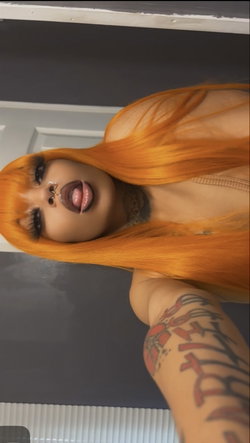 Woman with vibrant orange hair, visible tattoos on neck and arm, wearing a sheer top, posing indoors with lips parted.