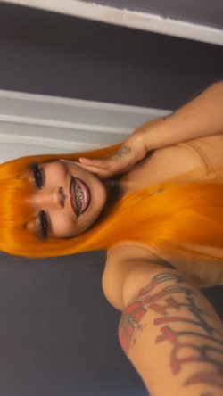 Woman with bright orange hair, visible tattoos on arm and hand, wearing a beige top, smiling in a close-up selfie