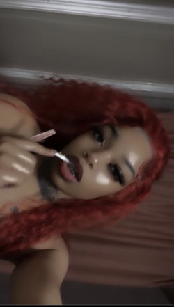 Woman with vibrant red curly hair and bold makeup, lying down while applying lip gloss, visible tattoos on shoulder and
