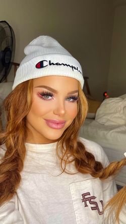 A woman with long wavy red hair wears a white Champion beanie and a white FSU sweatshirt, smiling in a bedroom setting.
