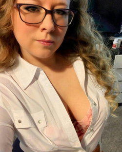 Blonde woman with curly hair and glasses wears an unbuttoned white shirt revealing pink lace bra, taking a close-up