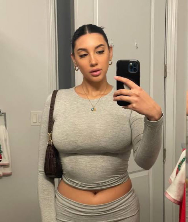 Curvy woman in a gray long-sleeve crop top and matching skirt takes a mirror selfie in a bathroom, holding a phone.