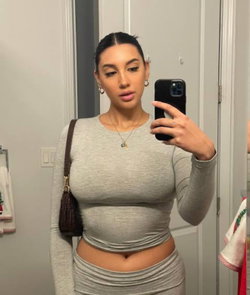 Curvy woman in a gray long-sleeve crop top and matching skirt takes a mirror selfie in a bathroom, holding a phone.