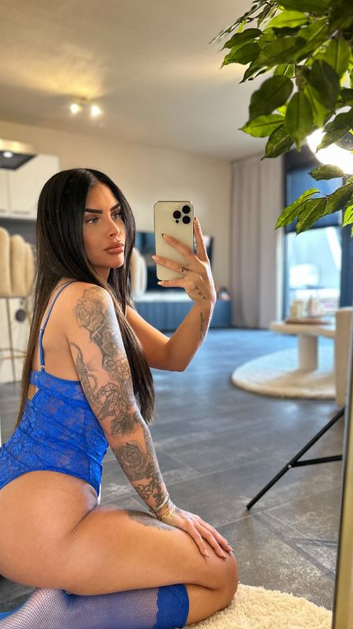 Curvy brunette with arm tattoos in blue lace bodysuit and fishnets kneels on rug in living room.
