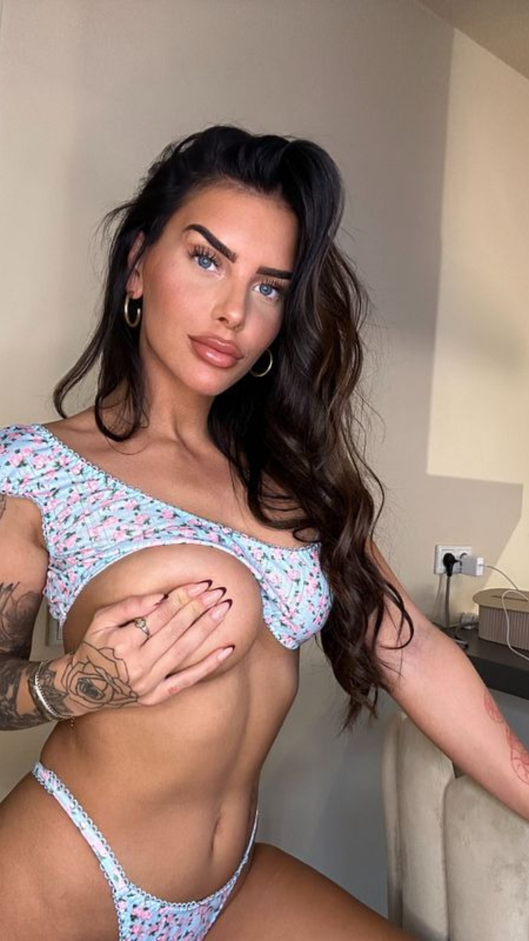 A curvy brunette with long dark hair and tattoos poses in a floral outfit indoors.