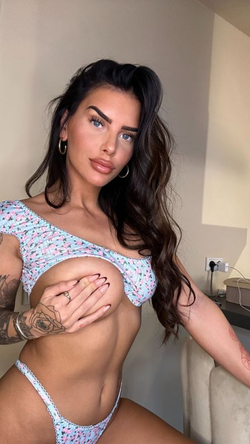 A curvy brunette with long dark hair and tattoos poses in a floral outfit indoors.