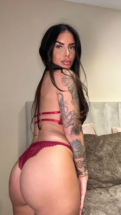 A curvy woman with long dark hair and tattoos wears red lingerie while posing in a bedroom.