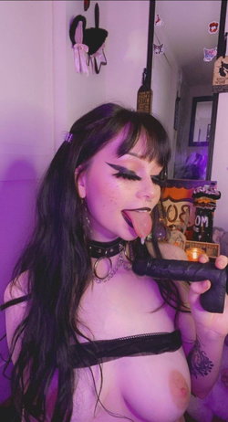 Brunette woman with long hair and bangs, wearing a black choker and sheer top, holding a toy while posing in a room