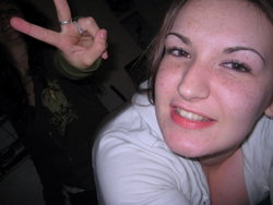 Freckled woman in a white hoodie smiles while taking a selfie with a peace sign indoors.