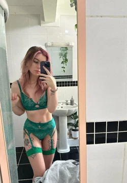 A woman with pink-tipped hair takes a mirror selfie in a green lace lingerie set, showing tattoos on her arms and