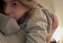 Blonde woman in a gray hoodie and blue lace underwear, lying on her side with head resting on arm, looking at camera in