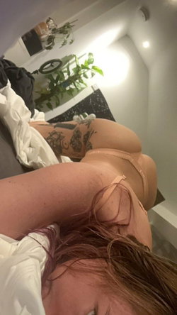 Curvy woman with tattoos on her legs lying on a bed, wearing peach-colored lingerie, in a dimly lit room with plants