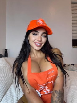 Woman with long dark hair wearing an orange cap and matching apron, smiling at the camera while seated on a bed.