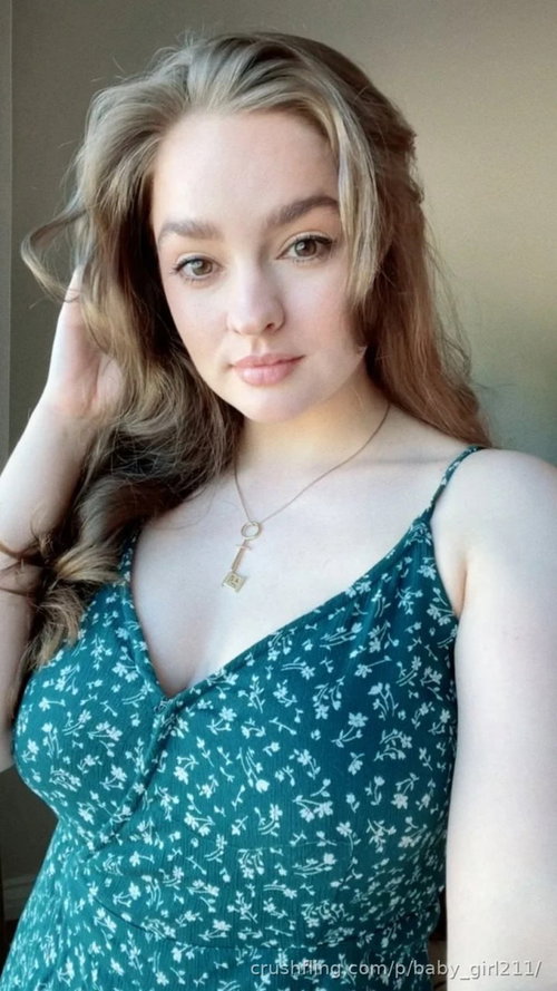 A woman with wavy blonde hair wears a teal floral spaghetti strap top, posing indoors with one hand in her hair.