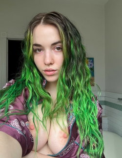 Person with long green hair in a bathroom, wearing a purple top with a pattern.