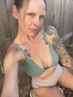 Woman with tattoos and blonde hair in a green bikini top and white shorts sitting outdoors.