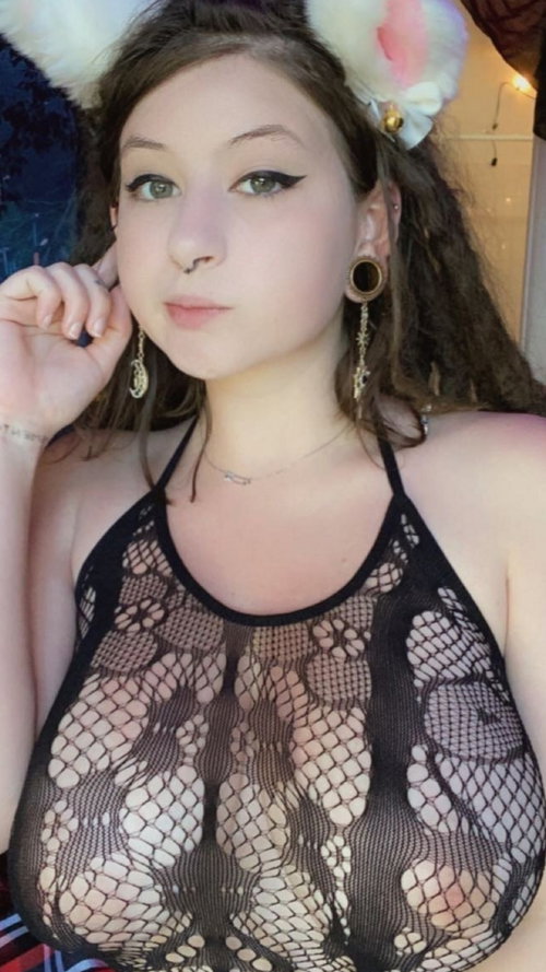 Brunette woman in a black mesh top with bunny ears, posing indoors with hand near face.