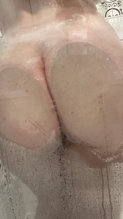 Close-up of a person's bare buttocks in a shower with soap suds, viewed through a glass door.